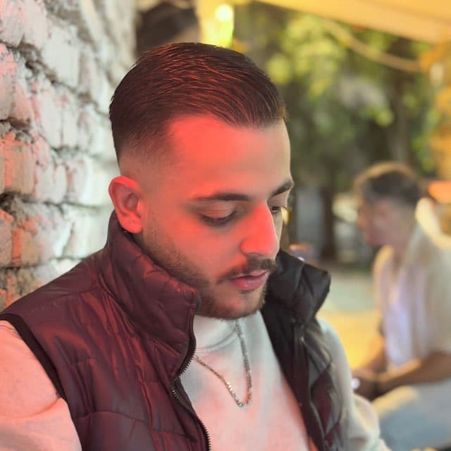 Mahdi Moosavi looking down, leaning against a stone wall outdoors in warm evening light, wearing a burgundy quilted vest.